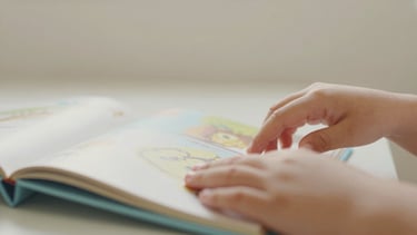 Close-up of a child's hands gently turning pages of a picture book in a soft-lit reading corner, natural light, candid, shallow depth of field.