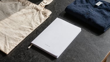 Flat-lay arrangement of branded merchandise — tote bag, printed notebook, and folded apparel on dark matte surface under north-facing daylight, showing print registration and fabric texture at close range