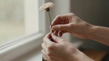 Close-up of hands gently arranging a single delicate flower stem into a small vase, soft natural window light, organic earthy tones, shallow depth of field, 35mm film