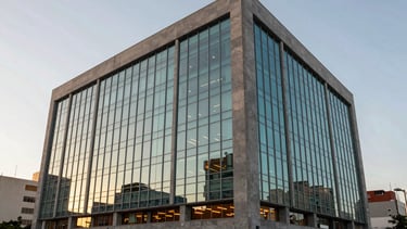 Professional photography of a modern, high-tech corporate office building in a South American/Brazilian business district. The shot is taken during the golden hour with soft sunlight. The architecture features clean glass panels and grey stone, with subtle cyan blue reflections and orange interior lighting visible through the windows. The composition is wide-angle and clean.
