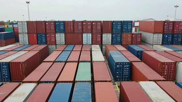 Wide overhead view of organized shipping containers stacked in precise rows at a container terminal, shot in bright overcast daylight for even exposure, emphasizing scale and mechanical grid-like arrangement, no people visible