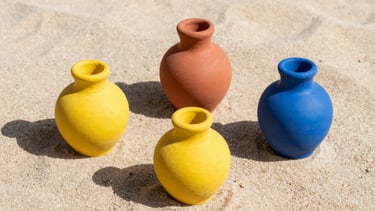 Three sculptural ceramic objects with highly saturated colors of yellow, blue, and terracotta, arranged on a sand-textured surface under sharp direct midday sun casting long dramatic shadows, 35mm lens