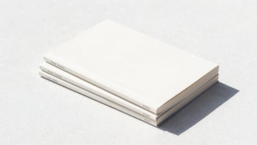 Three minimalist book covers stacked neatly on a clean white concrete surface, bright high-contrast daylight, 35mm
