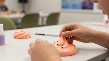 A close-up, high-quality photograph of a premium slime workshop for kids in a Southern European event space. In the center, a pair of hands are decorating a vibrant, peach-colored slime with elegant glitter and charm beads. The background is softly blurred, showing muted olive and off-white decor, reflecting a modern, creative, and professional animation setup.