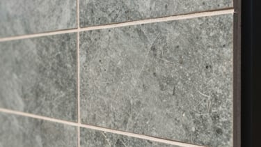 A close-up professional photograph of high-end architectural finishing in a South American / Brazilian modern house. The shot focuses on the precision of slate green and ash gray ceramic tile installation on a wall, showing perfect grout lines and clean edges under bright, natural daylight. The composition is sophisticated and minimalist.