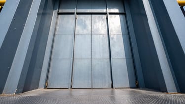 Low-angle architectural shot of a massive industrial freight elevator with open heavy steel doors, rugged textured steel floors, cool high-contrast daylight.