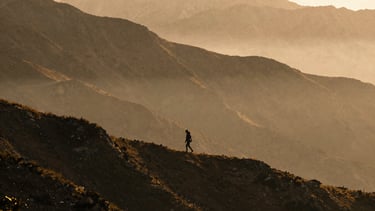 Wide environmental landscape of a solitary figure walking along a misty mountain ridge at golden hour, deep charcoal shadows and warm amber light, 35mm grain.