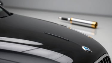 Macro photography of a black luxury car hood showing a perfect mirror reflection of a modern, minimalist studio. A professional detailing tool with silver and gold accents sits on a clean work surface in the background. Western European / French aesthetic, high-end automotive care.