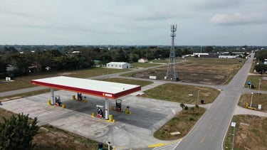 Wide drone overhead shot, overcast daylight, a completed fuel station canopy and pump islands at left, a three-legged cell tower visible in the mid-ground right, open lot and access road showing full site footprint and landscape relationship