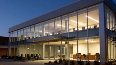 A wide shot of a North American tech hub office at dusk. The warm interior lighting contrasts with the deep navy evening light outside. The scene is efficient, professional, and sophisticated.