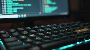 Macro close-up of a mechanical keyboard under low ambient neon-cyan light, with a blurred monitor in the background displaying terminal telemetry streams, shallow depth of field.