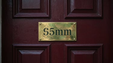 High-contrast close-up of an ornate, dark wood door and brass plaque of a historic financial institution, soft directional light highlighting texture. Editorial heritage photography, soft natural window light, muted deep crimson and forest green tones, 35mm, shallow depth of field.