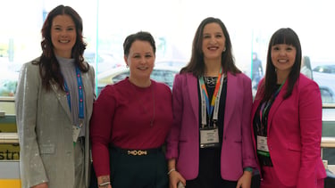 Four professional women standing by a window with a city view, wearing name tag lanyards and smiling for a group photo.