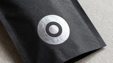Detail shot of a minimalist product package. Matte black cardstock with a silver embossed logo. Soft grey shadows on an off-white stone surface. Premium feel.