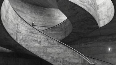 A black and white architectural shot of a spiral concrete staircase in a modern museum. Cinematic shadows and highlights. Dramatic perspective. Western European minimalist style.