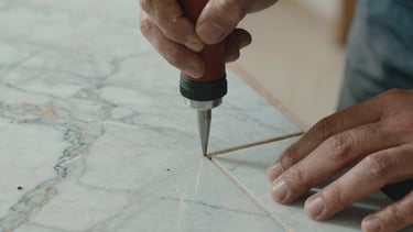 Close-up photography of a professional craftsman in Europe Centrale / France, using a precision suction tool to lay a massive marble-effect porcelain slab. The lighting is soft and natural, highlighting the matte texture of the tile and the meticulous grout alignment. Muted Blue and Warm Brown tones dominate the palette.