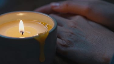 Macro close-up of warm golden wax pooling in a dark ceramic vessel, low-key candlelight casting deep blue shadows, soft skin contact in the background
