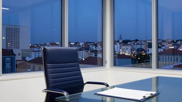 A high-end law office interior in Lisbon, European Portuguese setting. A view of a designer chair and a modern glass table with a single file folder. Large windows show the Lisbon skyline at dusk. Professional, trustworthy, and modern atmosphere. Dominant colors are navy blue and white.
