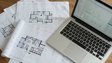 Top-down structured desk layout showing architectural blueprints next to a modern laptop screen displaying data charts, crisp daylight.