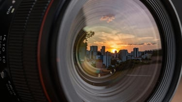 Close-up of a high-end camera lens reflecting a sunset scene in a Brazilian city. The image highlights the intersection of raw capture and technical finesse.