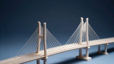 A minimalist studio shot of an architectural model representing a bridge, symbolizing the infrastructure for payments. Lighting is dramatic with deep navy and pale blue highlights.