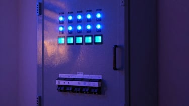 Professional photography of a high-tech electrical control panel in a modern South American home, with glowing blue and cyan status lights, reflecting a secure and advanced technological environment, dark blue and purple ambient lighting.