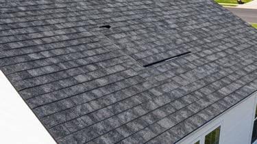 A professional high-angle photograph of a newly installed charcoal gray asphalt shingle roof on a modern suburban North American home. The sun is shining brightly, showing the crisp edges and quality craftsmanship. The house has clean white siding and the scene is set in a well-manicured New Jersey neighborhood.