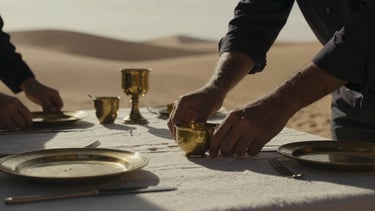 An intimate close-up of hands arranging a private luxury camp table in the Moroccan desert, heavy shadows, soft overcast daylight, rich textures of linen and brass