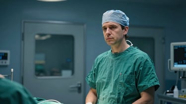 Cinematic over-the-shoulder shot of a European surgeon in green scrubs standing before the sterile operating room doors, cool clinical lighting.