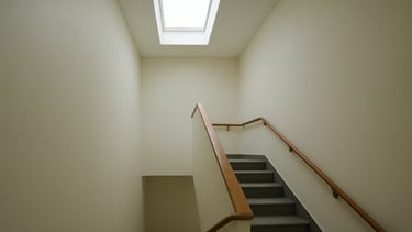 Wide panoramic view of a freshly painted residential stairwell in a multi-storey building, natural overcast light from skylight above, smooth off-white walls on all sides, clean paint line at handrail, no people, documentary style