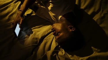 A man lying in bed at night using a glowing smartphone with blue light in a dark room.