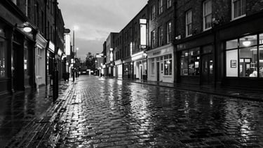 Panoramic 21:9 frame, high contrast black and white, gritty urban Birmingham Jewellery Quarter street at dusk, wet cobblestones reflecting neon signs, wide-angle perspective, low horizon.