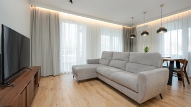 Nowoczesny salon z jasną narożną sofą, telewizorem, drewnianą podłogą i zasłonami.