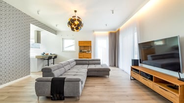 Nowoczesny salon z szarą narożną sofą, telewizorem na drewnianej szafce i aneksem kuchennym.