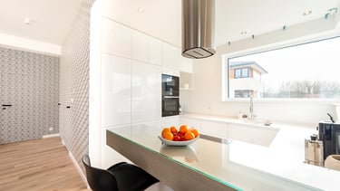 Biała kuchnia z wyspą, czarnymi hokerami i miską owoców na blacie. Drzwi ukryte, tapeta