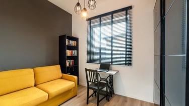 Pokój z żółtą sofą, biurkiem z laptopem przy oknie z żaluzjami i regałem.