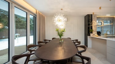 Nowoczesna jadalnia z długim stołem, krzesłami, lampą sufitową i kuchnią po prawej.