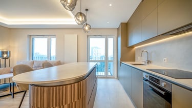 Kuchnia z wyspą, zabudową AGD, oświetleniem sufitowym oraz dużymi oknami balkonowymi.