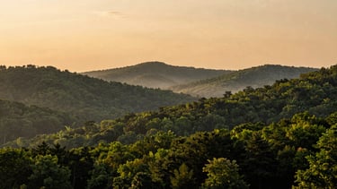 Scenic panoramic landscape of Cherokee County hills at golden hour, deep green forests, soft mist rising from the valleys, warm clay-toned horizon