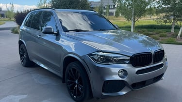 A silver BMW X5 luxury SUV with black rims parked on a driveway under a blue sky.