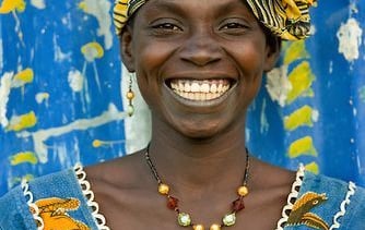 Smiling woman wearing a yellow African headwrap and patterned blue dress against a blue background.