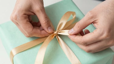 Close-up of hands carefully tying a warm gold ribbon around a mint green gift package, soft natural light