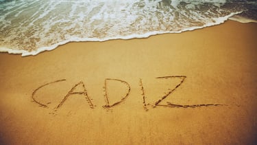 Foto de "Cádiz" escrito na areia da praia, com as ondas do mar ao fundo.
