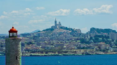 Vista do mar de Marselha com a basílica Notre-Dame de la Garde no topo e o farol na entrada do porto.