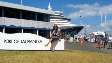 Foto da autora sentada no Porto de Tauranga, com um navio e a cidade ao fundo.