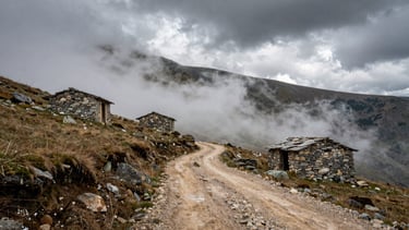 A high-altitude dirt path winding through the misty Kaçkar mountains, isolated stone shepherd huts, dramatic clouds.