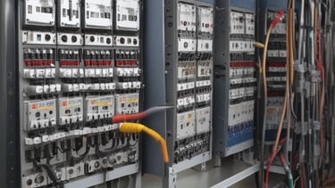 A construction site is depicted with exposed electrical wires protruding from a circular conduit in a rough concrete wall. The floor is covered in dust and debris, and there are multiple cables organized in a black mesh covering running along the floor.