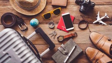 Travel essentials flat lay on a wooden table including suitcase, passport, camera, and hat.