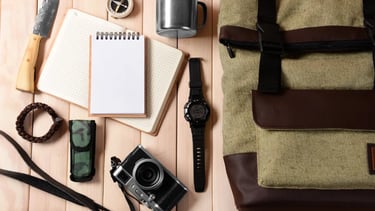 Survival gear and travel essentials including a canvas backpack, camera, notebook, and compass on wood.