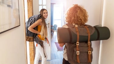 Two female backpackers with travel gear entering a bright hostel room for their vacation adventure.